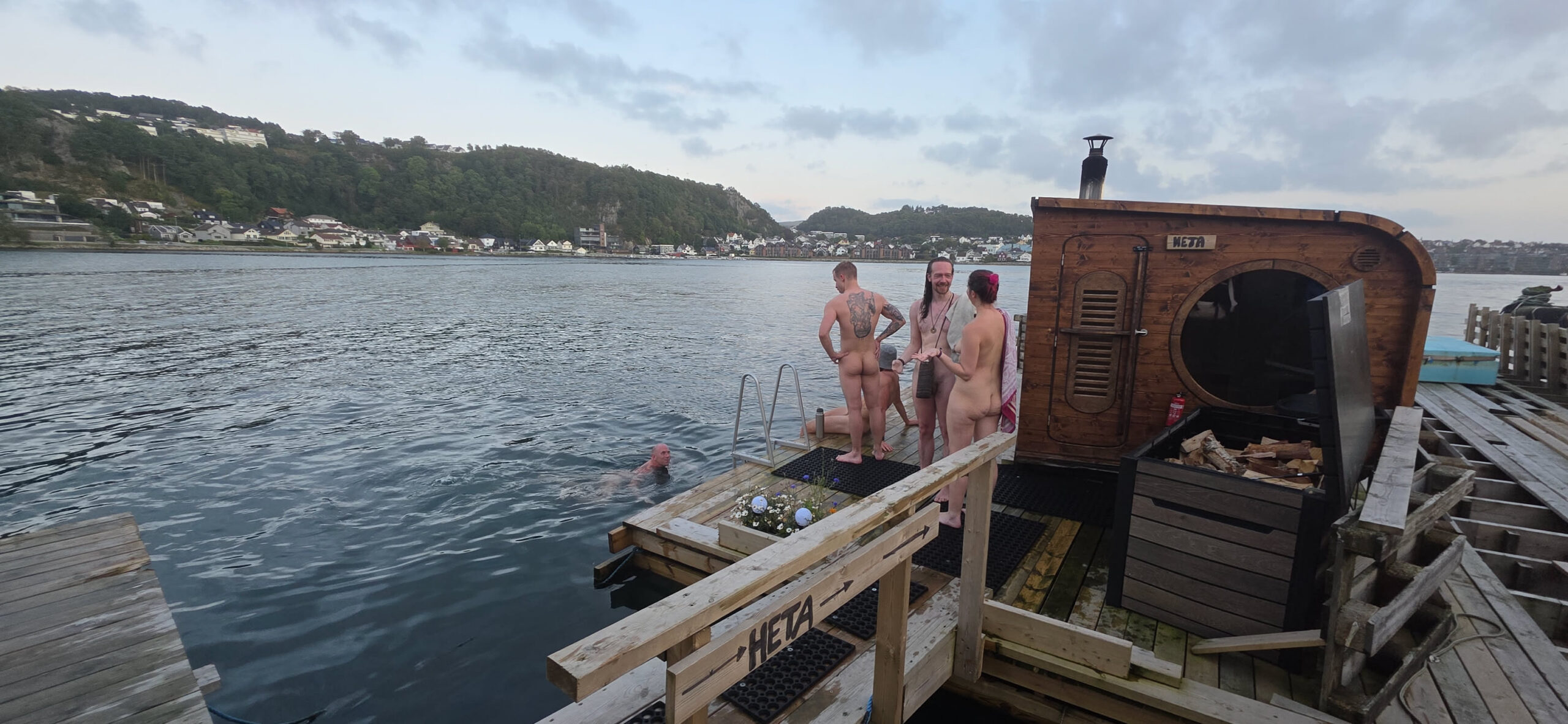 Mennesker utenfor badstu, med badende i Gandsfjorden