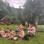 En gruppe nakne mennesker sitter ute i naturen og ser smilende mot kameraet.