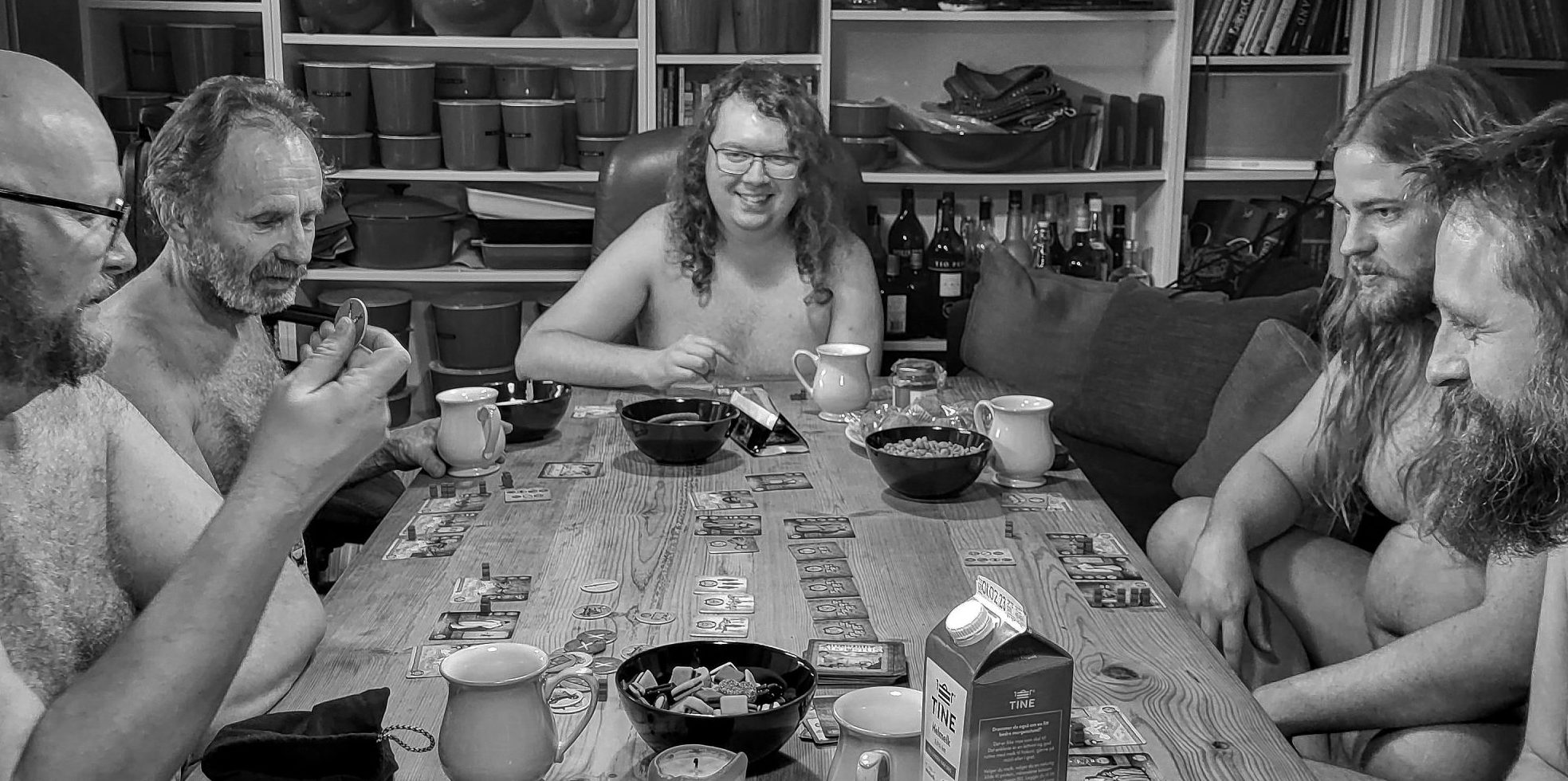 Fem nakne menn sitter rundt et kjøkkenbord og spiller brettspill